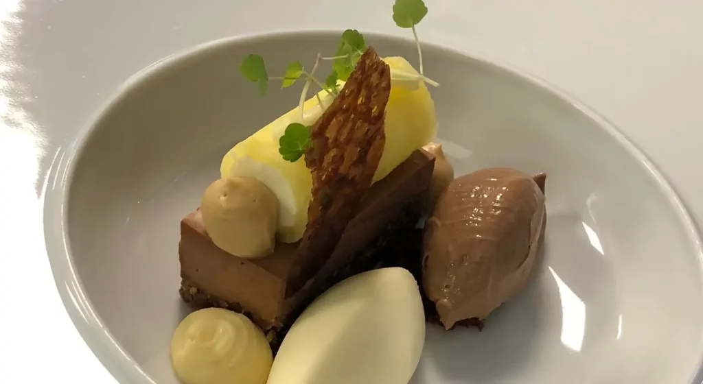 Een gastronomisch dessertbord met een plakje chocoladetaart, klodders room, een quenelle chocolademousse en gegarneerd met een knapperig wafeltje en kleine groene blaadjes voor privé