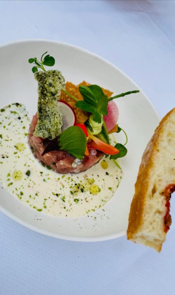 Een gastronomisch gerecht dat kunstig wordt gepresenteerd op een wit bord. Het bestaat uit in blokjes gesneden vlees, versierd met diverse groenten en kruiden, bovenop een romige saus. Een stuk brood met een zichtbare zongedroogde tomaat wordt gedeeltelijk weergegeven aan de rechterkant van het bord, klaar voor close-up in elke voedselmedia-functie.
