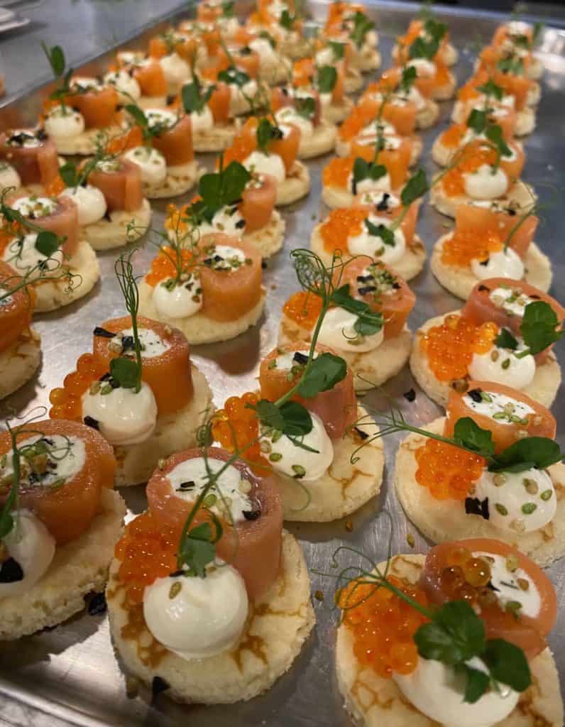 Een dienblad met prachtig gearrangeerde canapés met plakjes gerookte zalm, klodders roomkaas, oranje viskuiten en garnituren van verse kruiden op kleine, ronde bodems. De mediawaardige presentatie is elegant en smakelijk.