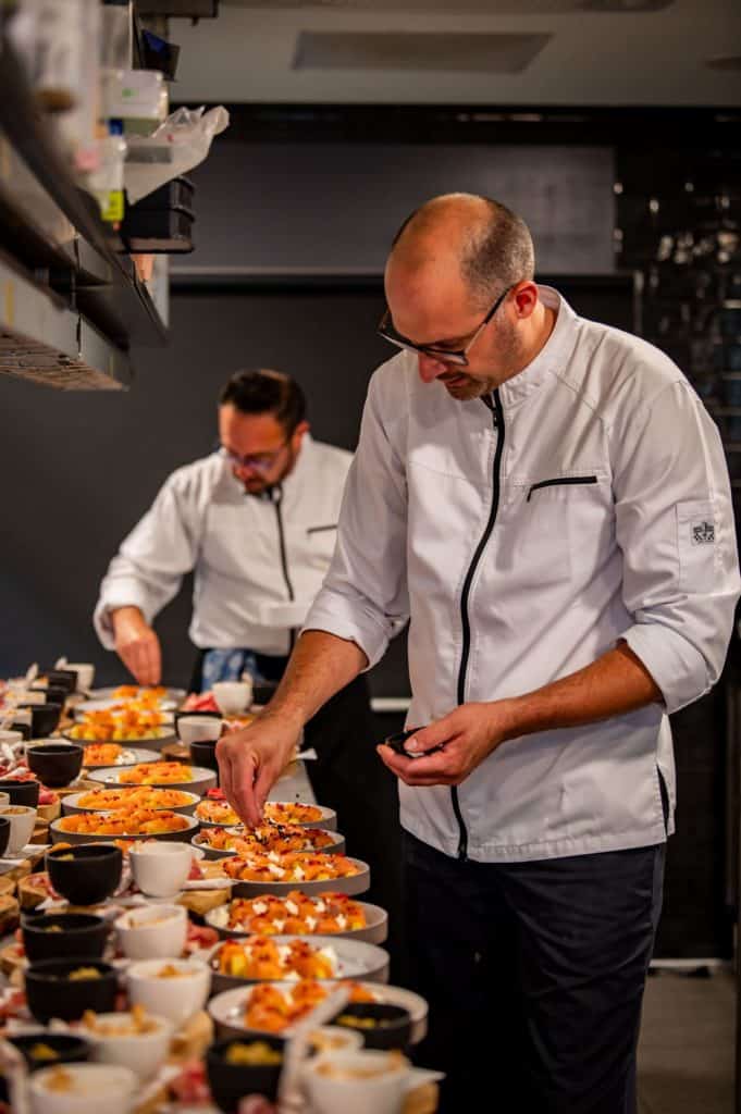 Twee in witte uniformen geklede chef-koks bereiden in een professionele keuken nauwgezet een verscheidenheid aan gerechten. Eén chef-kok aan de voorkant concentreert zich aandachtig op het serveren van gerechten, terwijl een andere chef-kok op de achtergrond ook werkt aan de presentatie van het eten, allemaal onder het toeziend oog van mediacamera's die elke beweging vastleggen.
