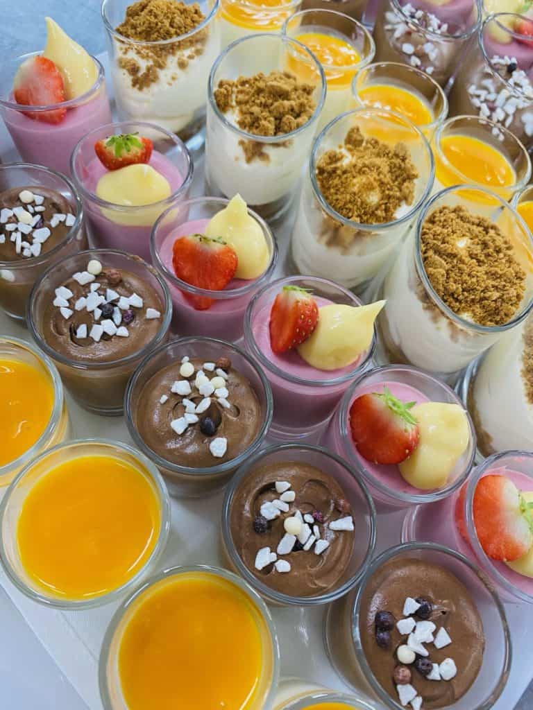 Een verscheidenheid aan kleurrijke dessertbekers op tafel, perfect voor elk media-evenement. De cups bevatten verschillende smaken waaronder chocolade, aardbei en citroen. Sommige zijn bedekt met gemalen koekjes, slagroom of verse aardbeien en gegarneerd met kleine decoratieve snoepjes.