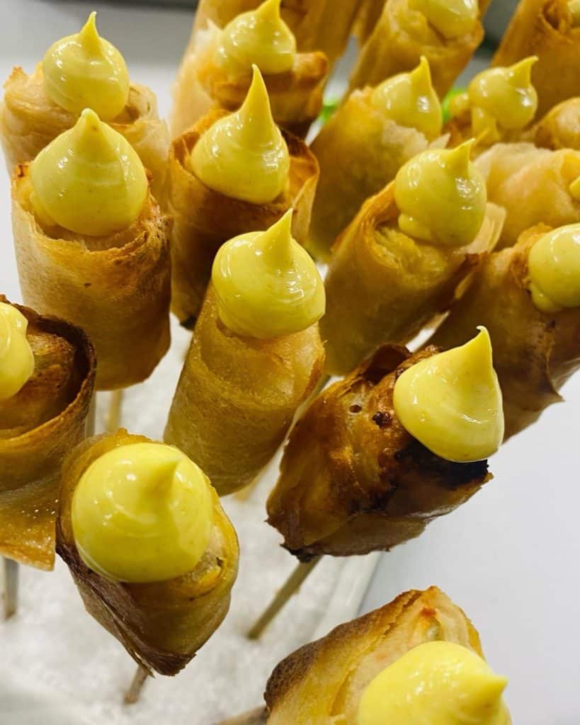 Close-up van knapperige loempiaspiesjes met daarop klodders felgele saus, rechtopstaand op een witte ondergrond. De broodjes zijn goudbruin, met de saus prominent op elk broodje, klaar om als voorgerecht of tussendoortje te worden geserveerd: een perfecte visuele traktatie voor je social media-feed.