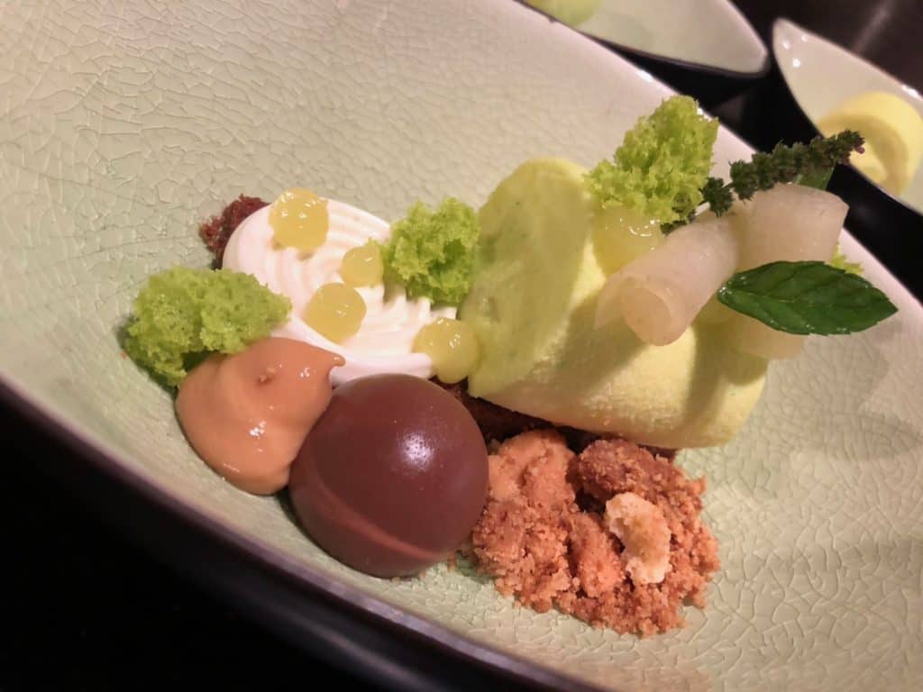 Een gastronomisch dessert, dat veel lof van de media verdient, wordt kunstig op een groen getextureerd bord geserveerd. Het bevat een cilindrische groene mousse, een klodder witte room, groene biscuitstukjes, karameldopjes, een chocoladebol, bruine brokjes en is gegarneerd met een muntblad en een klein takje.